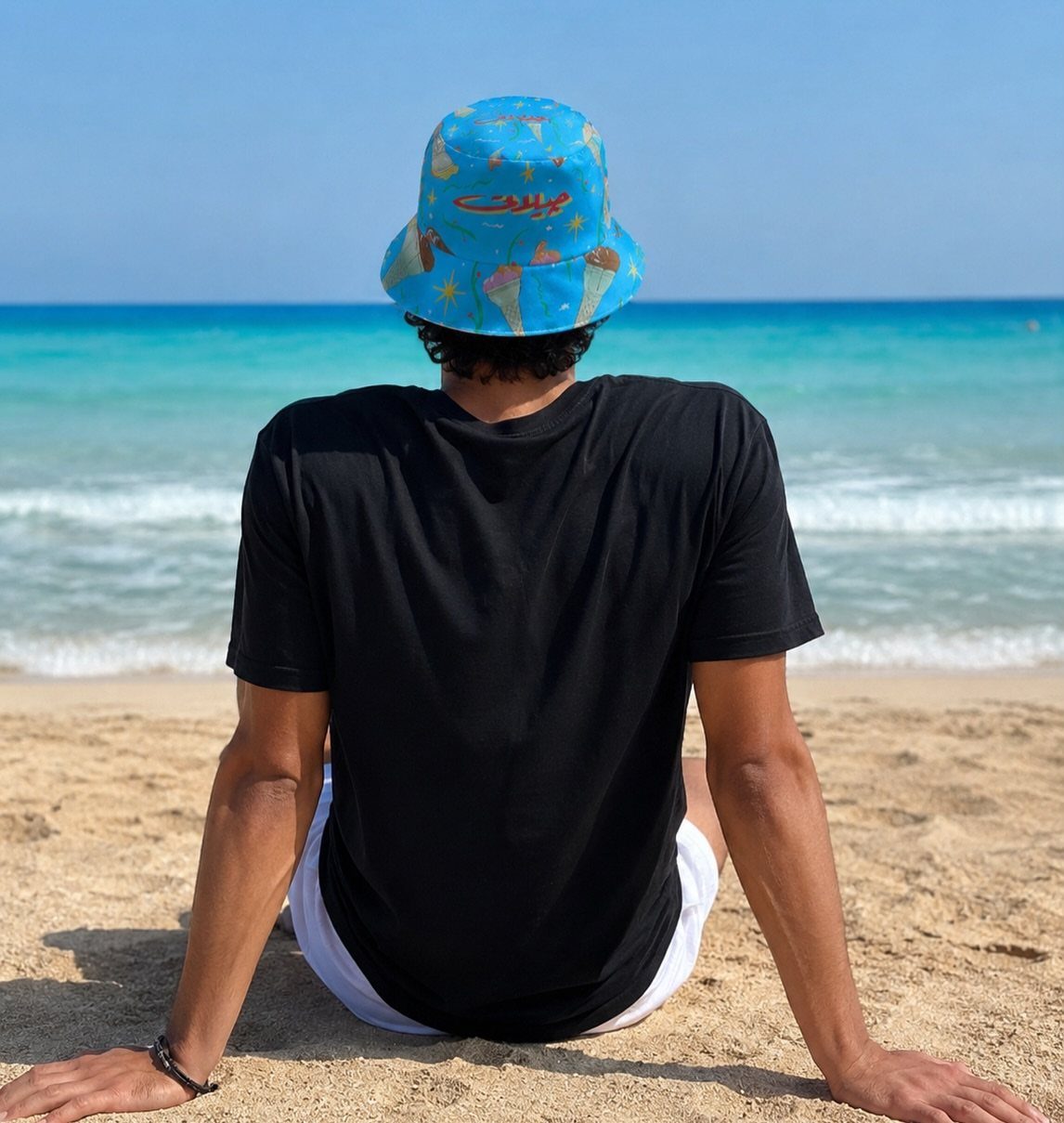 Man sitting on a beach wearing a blue patterned bucket hat and black t-shirt