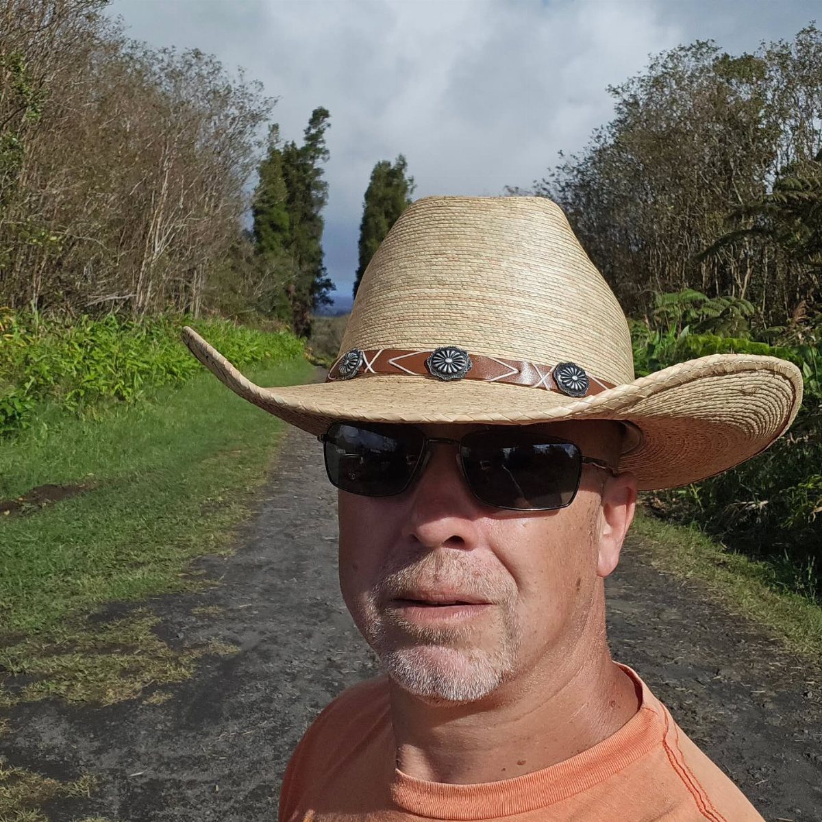 Man wearing a straw cowboy hat and sunglasses on a rural dirt path.