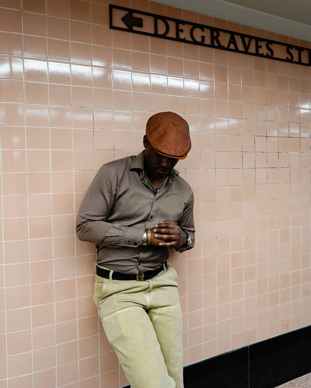 Man in a brown flat cap and striped shirt leaning against pink subway tiles.