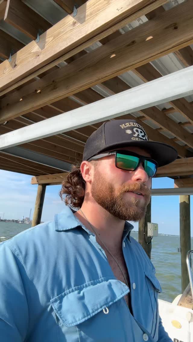 Man with beard wearing a black trucker hat and blue shirt by the water
