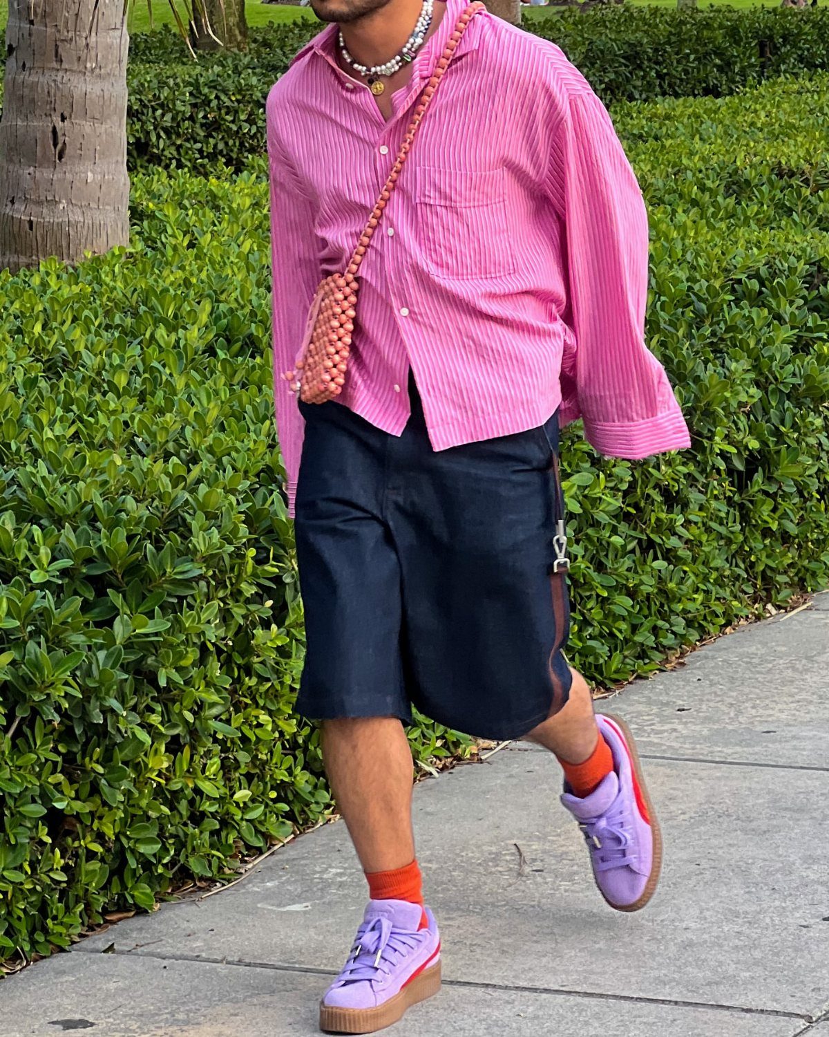 Man in pink striped shirt and navy shorts wearing a crossbody bag and necklace.