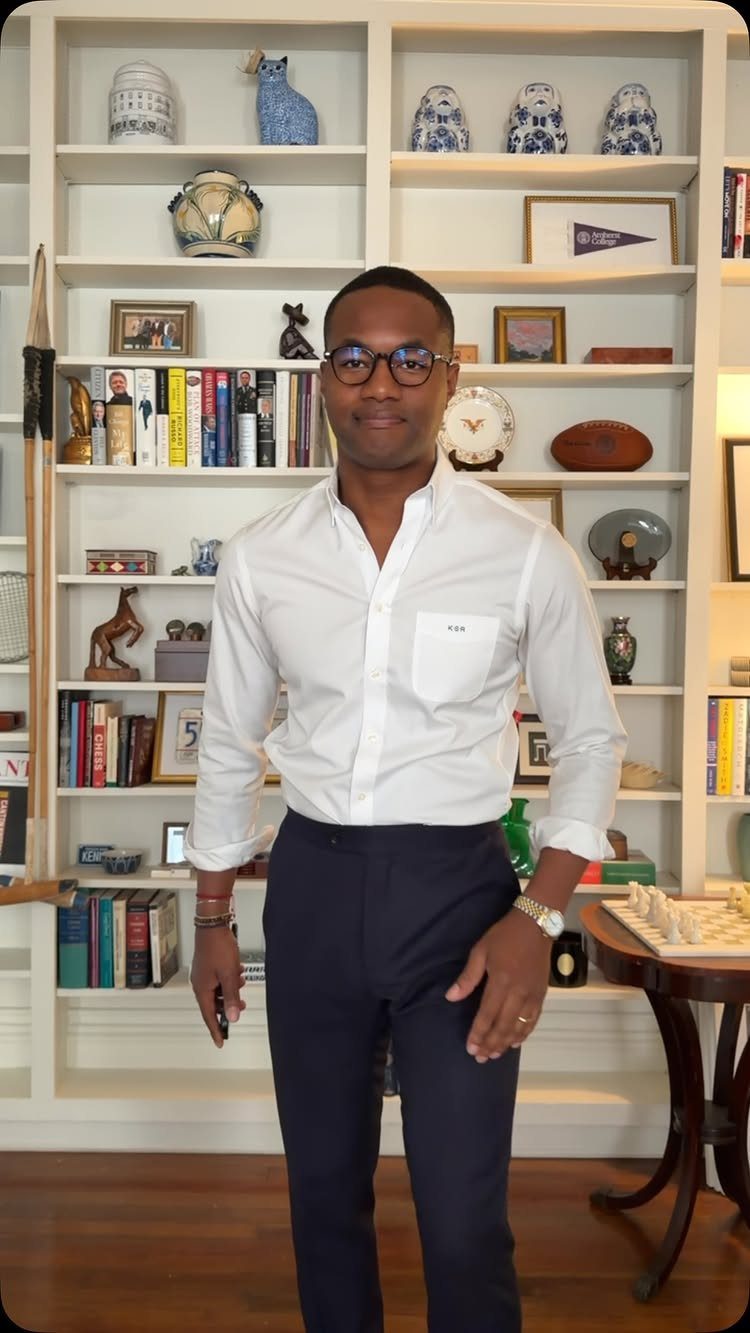 Man in white button-down shirt and navy trousers posing before a white bookshelf.