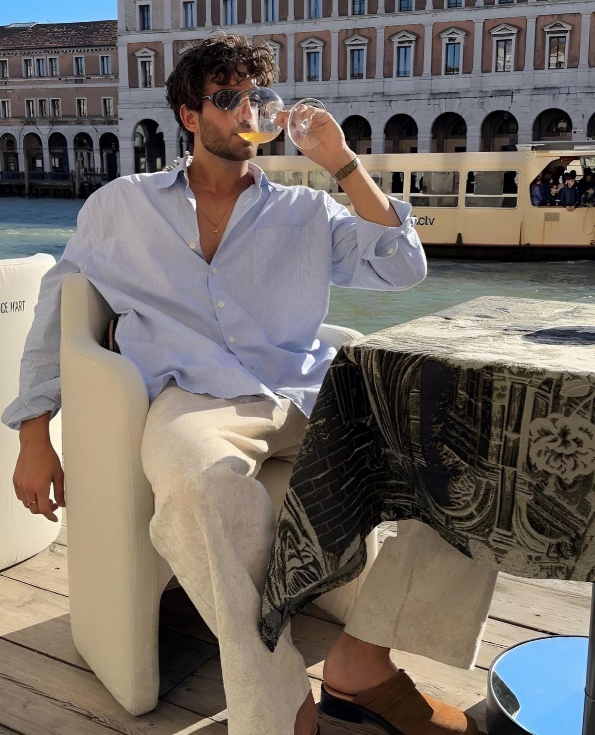 Man in light blue shirt and cream trousers drinking wine by a canal