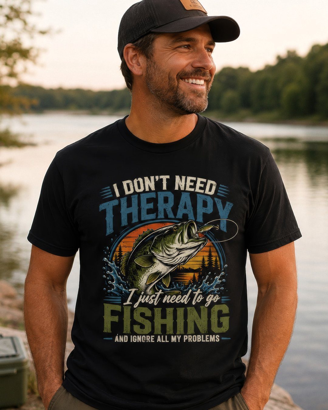 Man in black graphic t-shirt with fishing design and black cap by a river.