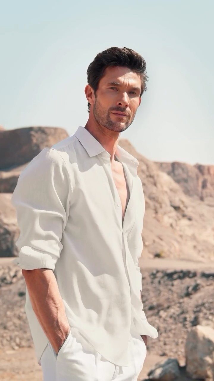 Man in a white button-down shirt and white trousers posing in a desert.