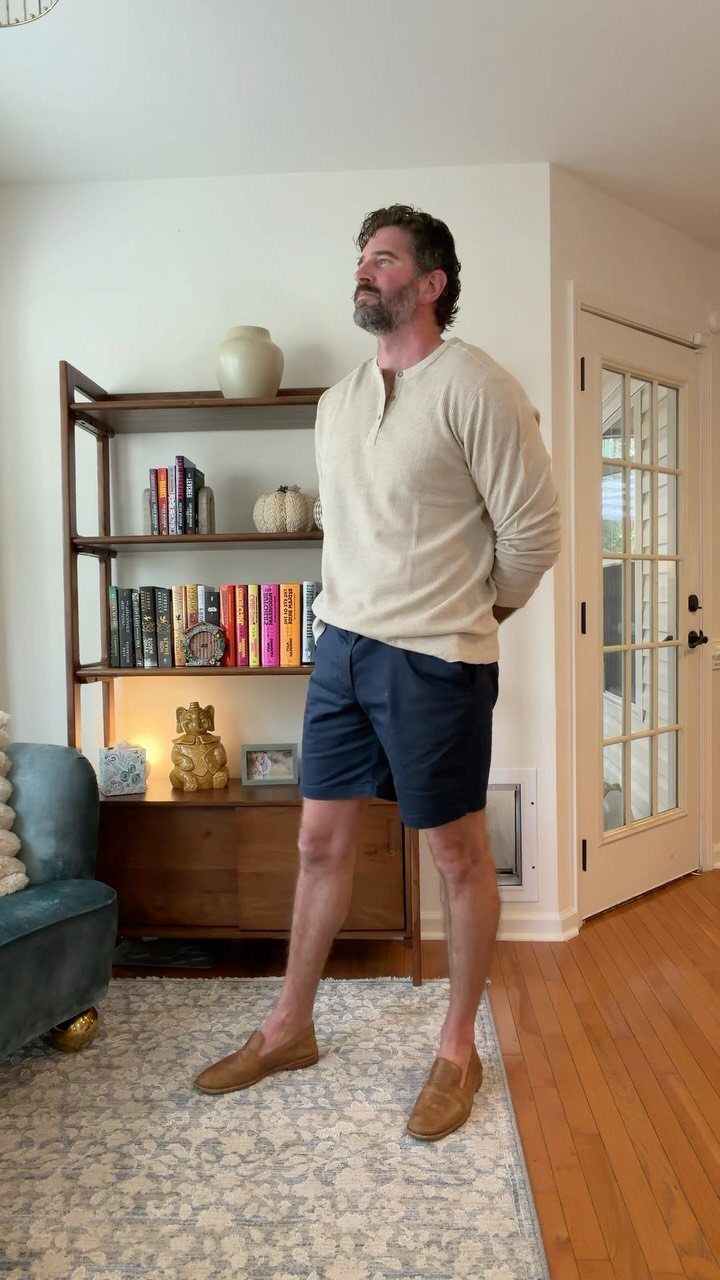 Man wearing a beige henley shirt, navy blue shorts, and brown loafers indoors.
