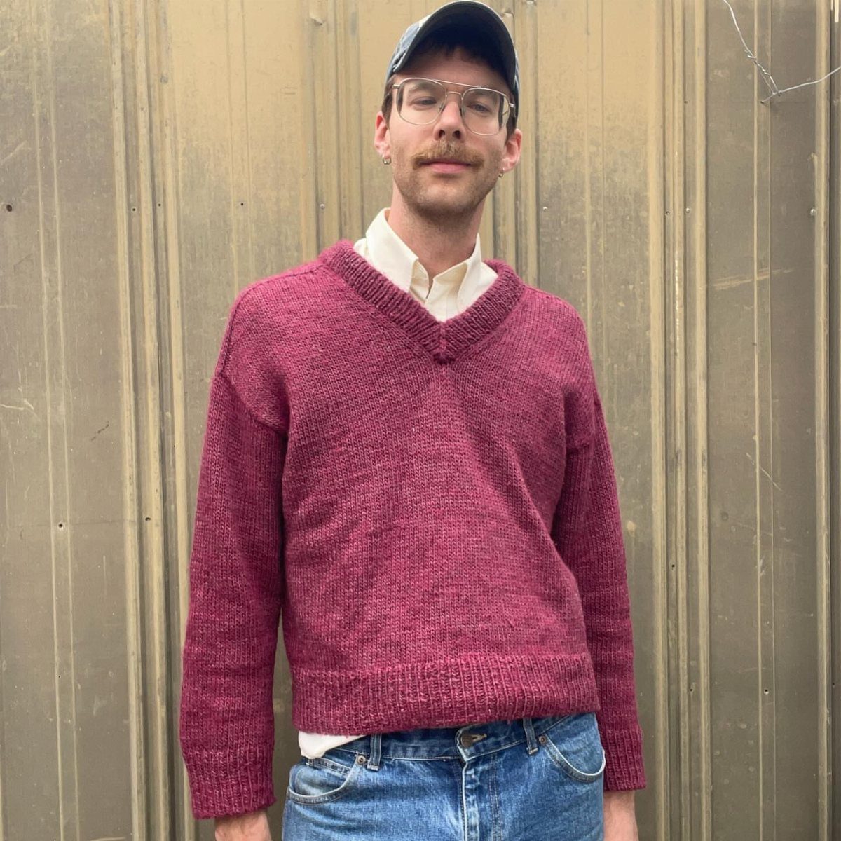 Man wearing a maroon v-neck knitted pullover, cream shirt, and blue jeans.