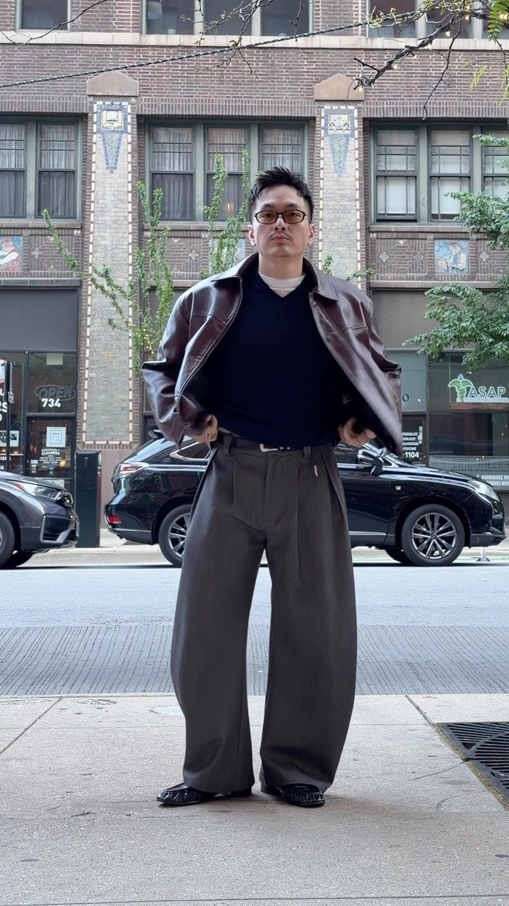 Man wearing a brown leather jacket over a navy sweater and wide grey trousers.