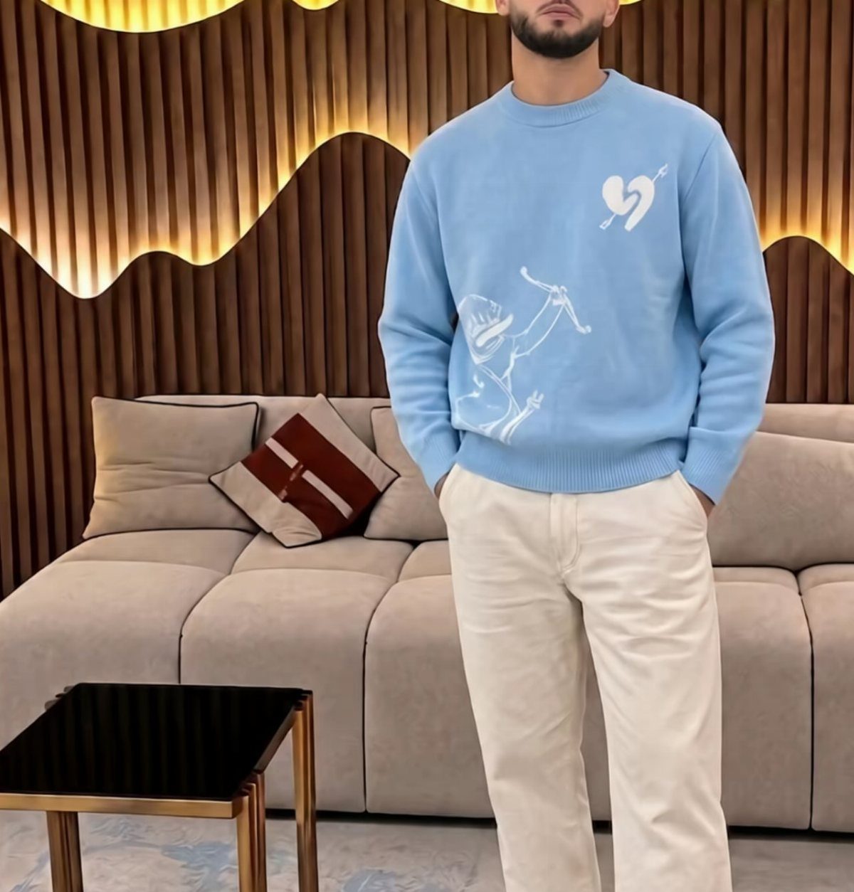 Man wearing a light blue Cupid sweatshirt and off-white trousers in a modern room.