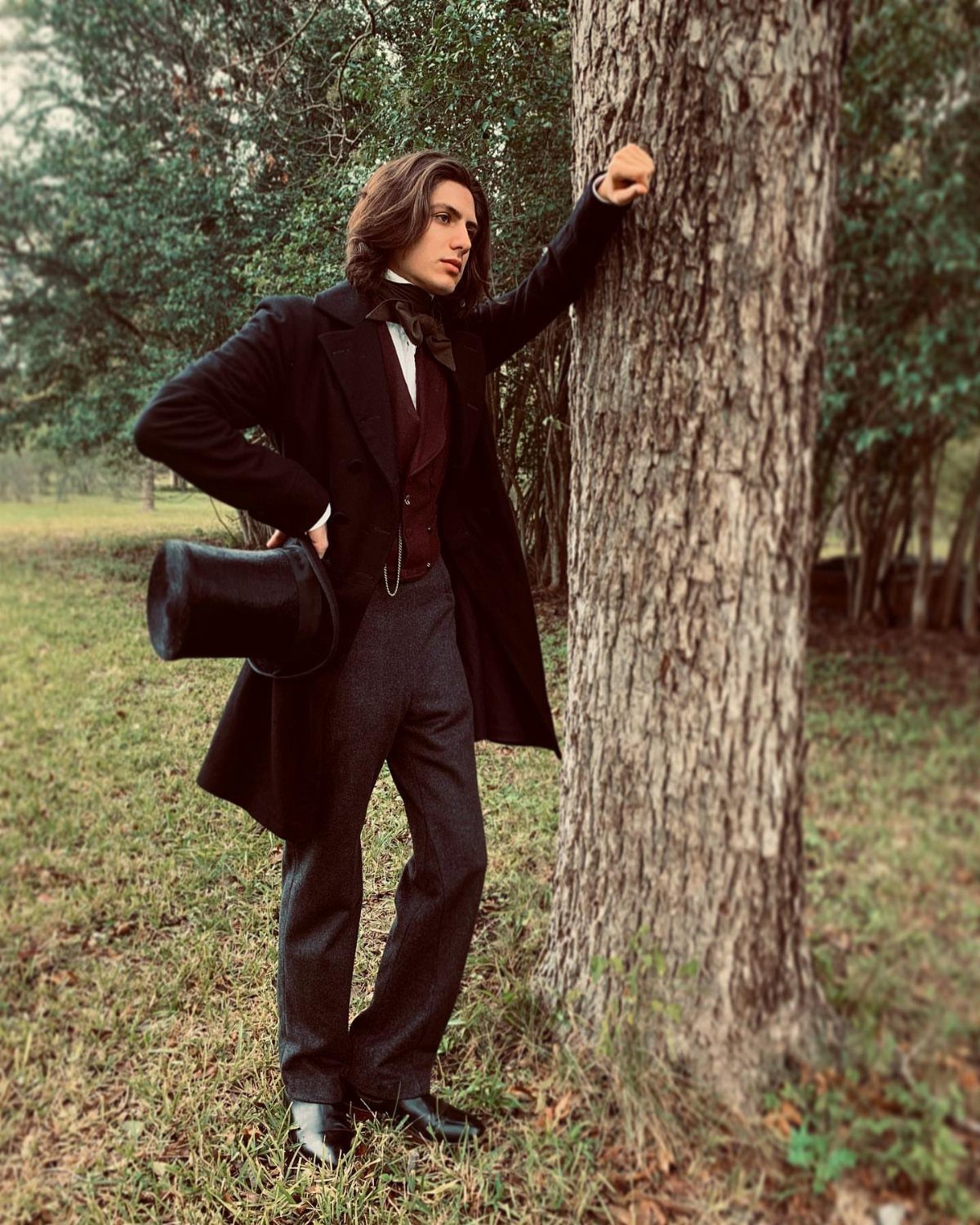 Man in Victorian attire leaning against a tree wearing a frock coat and waistcoat.