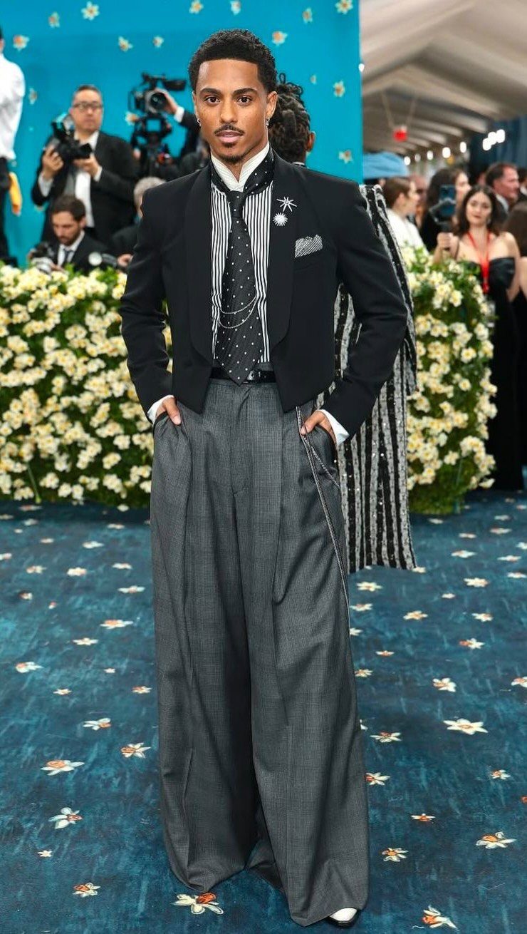 Man in a black cropped blazer and wide-leg grey trousers posing on blue carpet.