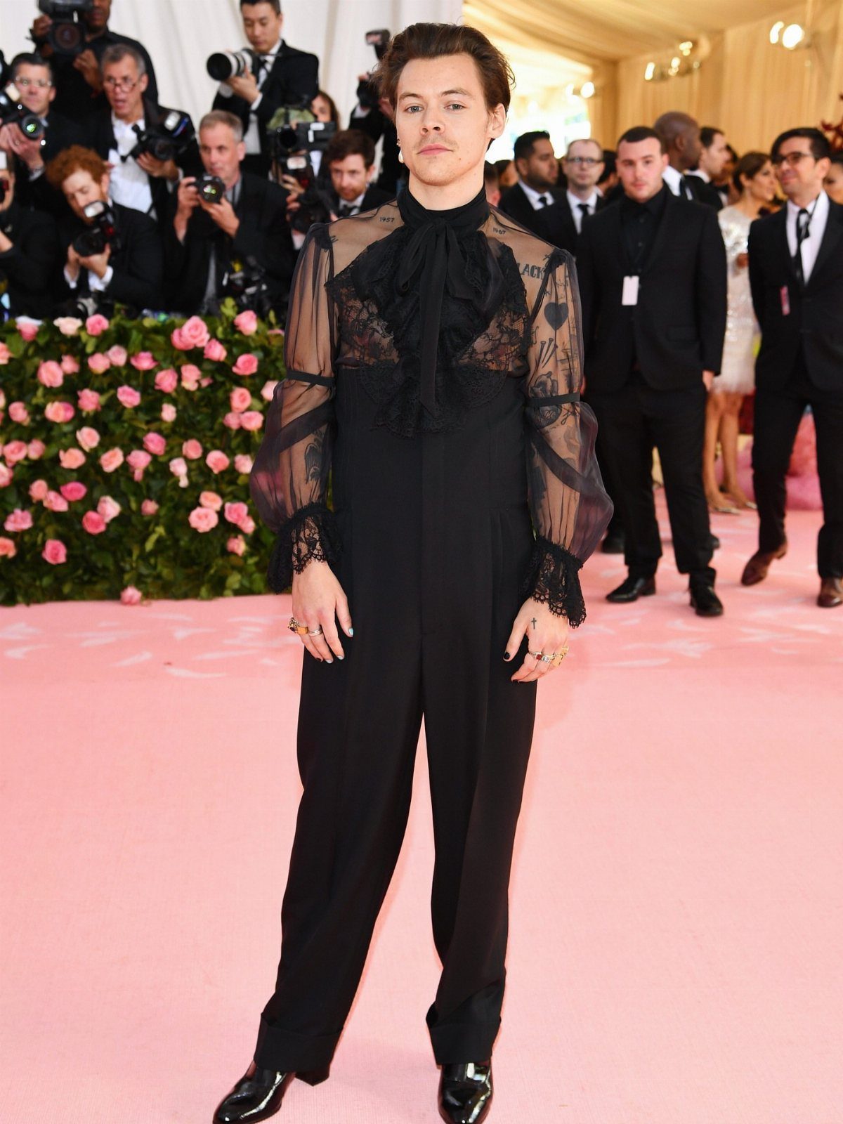 Harry Styles wearing a sheer black lace top and black trousers on pink carpet
