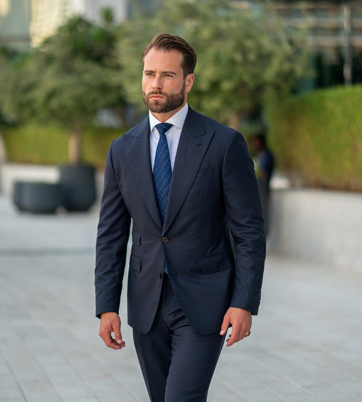 Man in a navy blue tailored suit and blue tie walking outdoors.