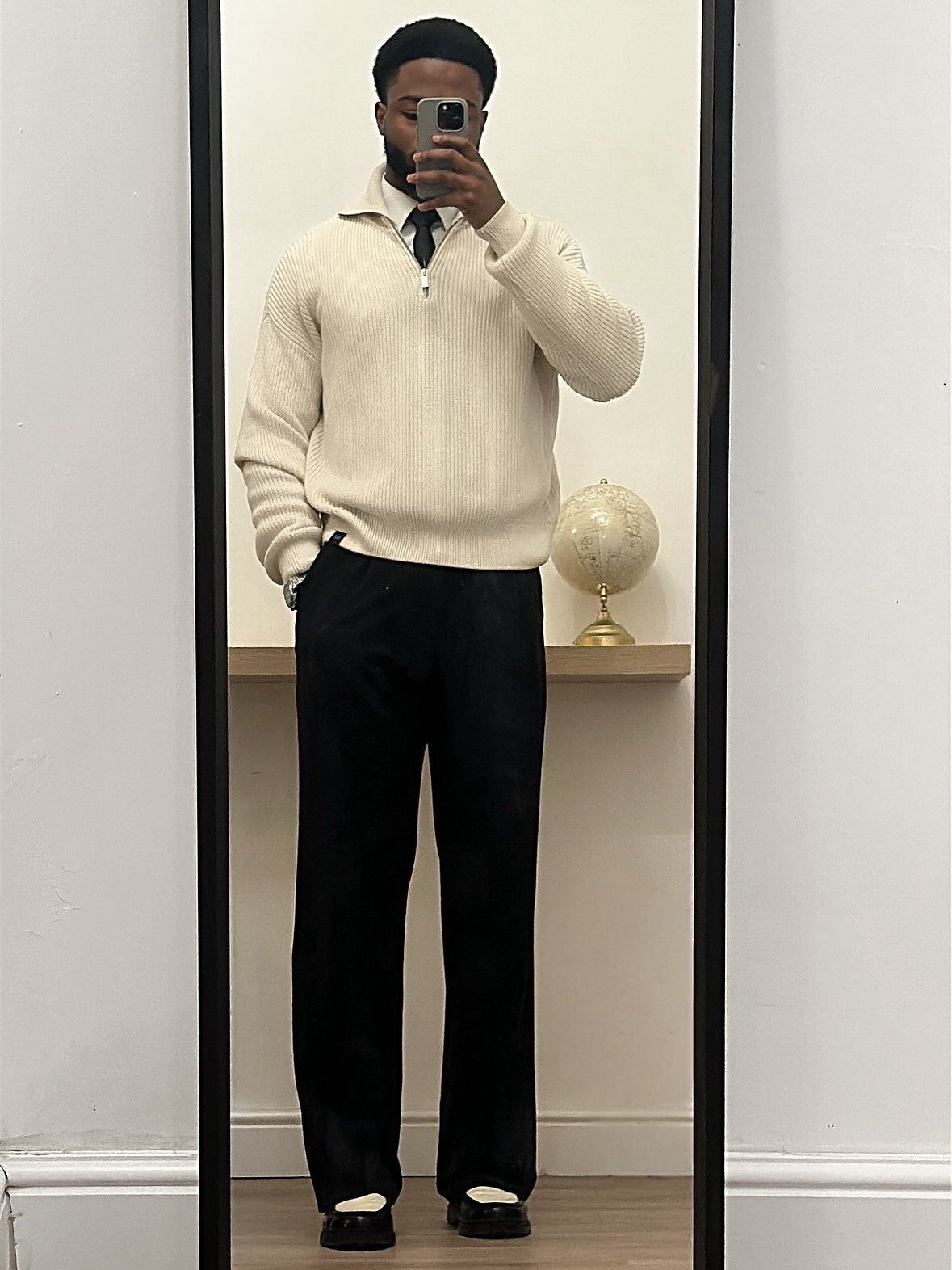 Man in cream ribbed quarter-zip sweater and black wide-leg trousers taking a mirror selfie.