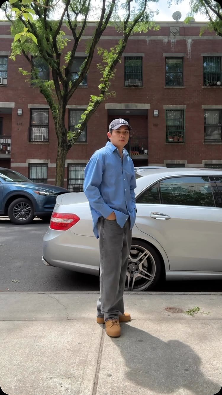 Man in light blue shirt and grey trousers standing by a silver car.