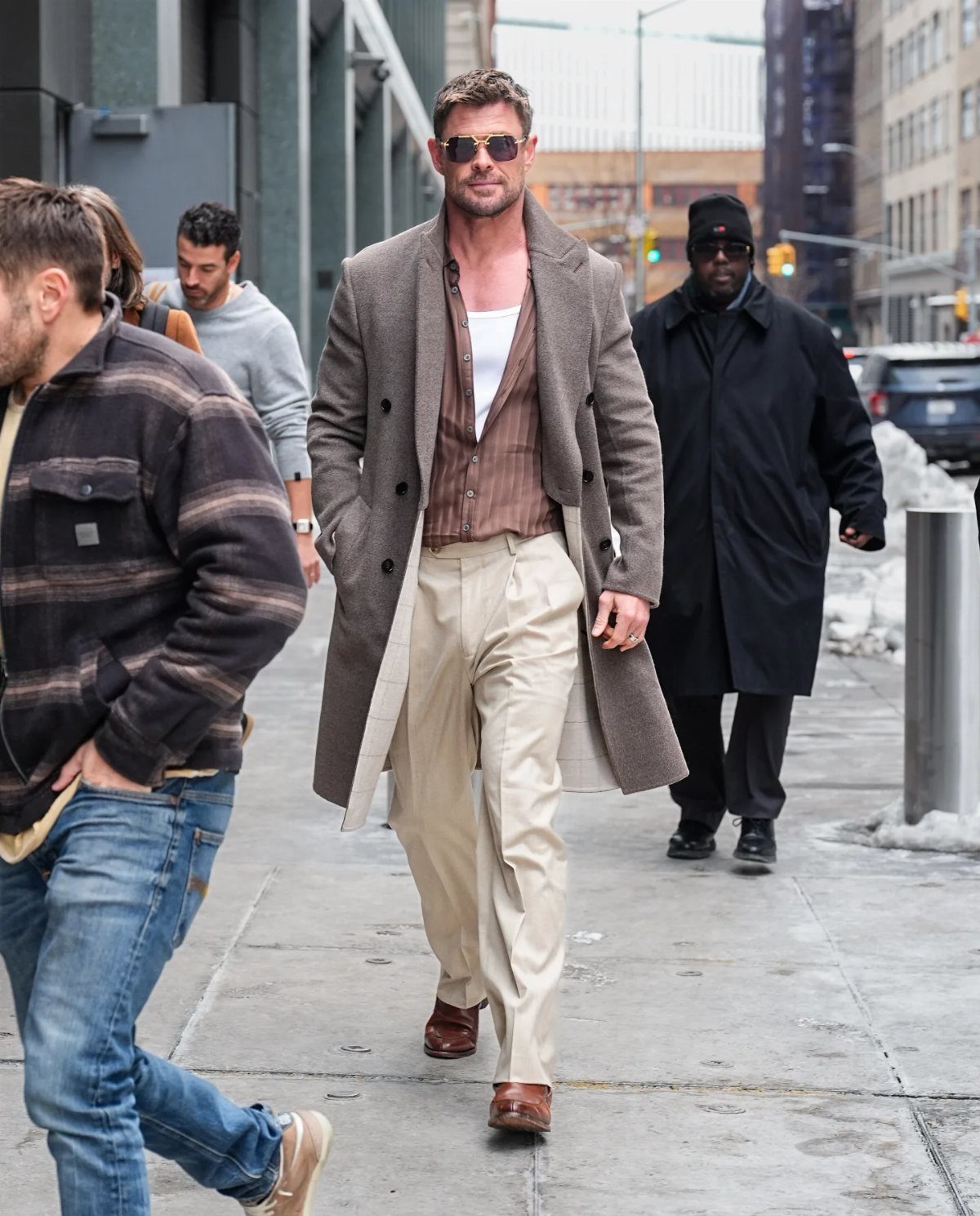 Man in a grey overcoat, brown shirt, and beige trousers walking on city street.