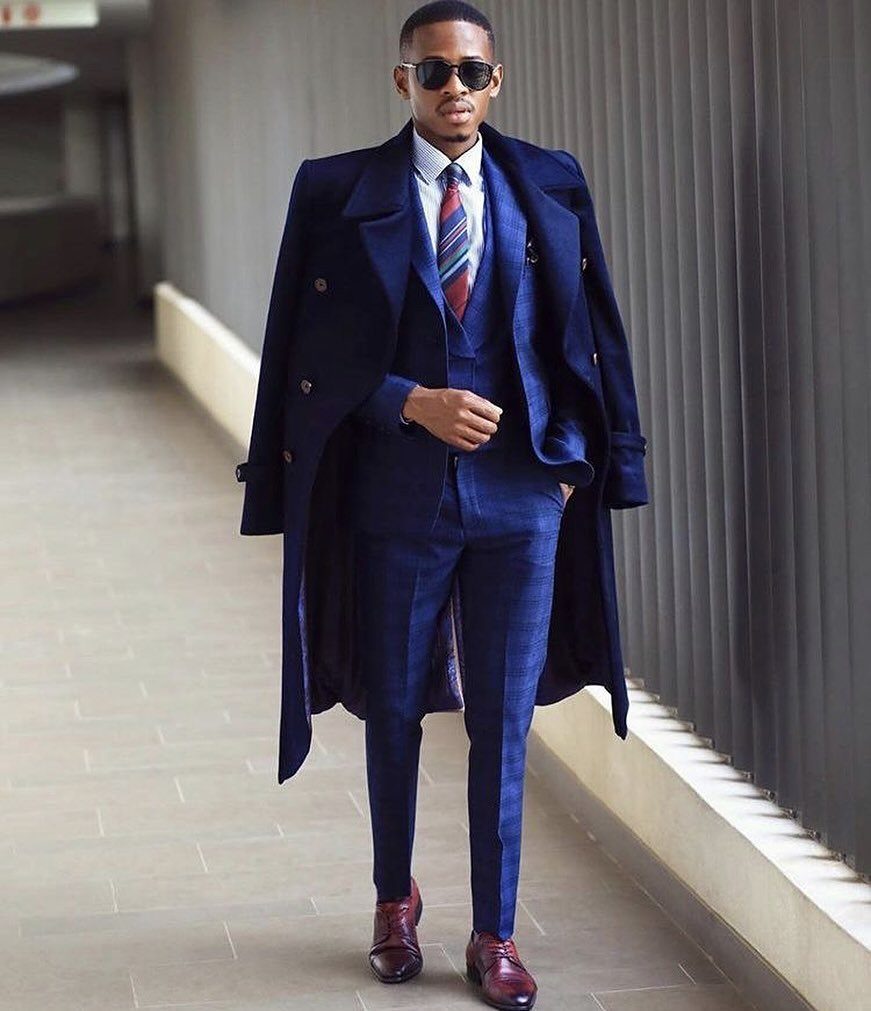 Man in navy blue three-piece suit and overcoat wearing burgundy dress shoes.