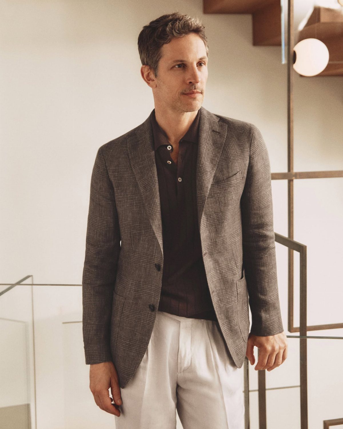 Man wearing a brown relaxed tailoring jacket, dark polo, and white trousers indoors.