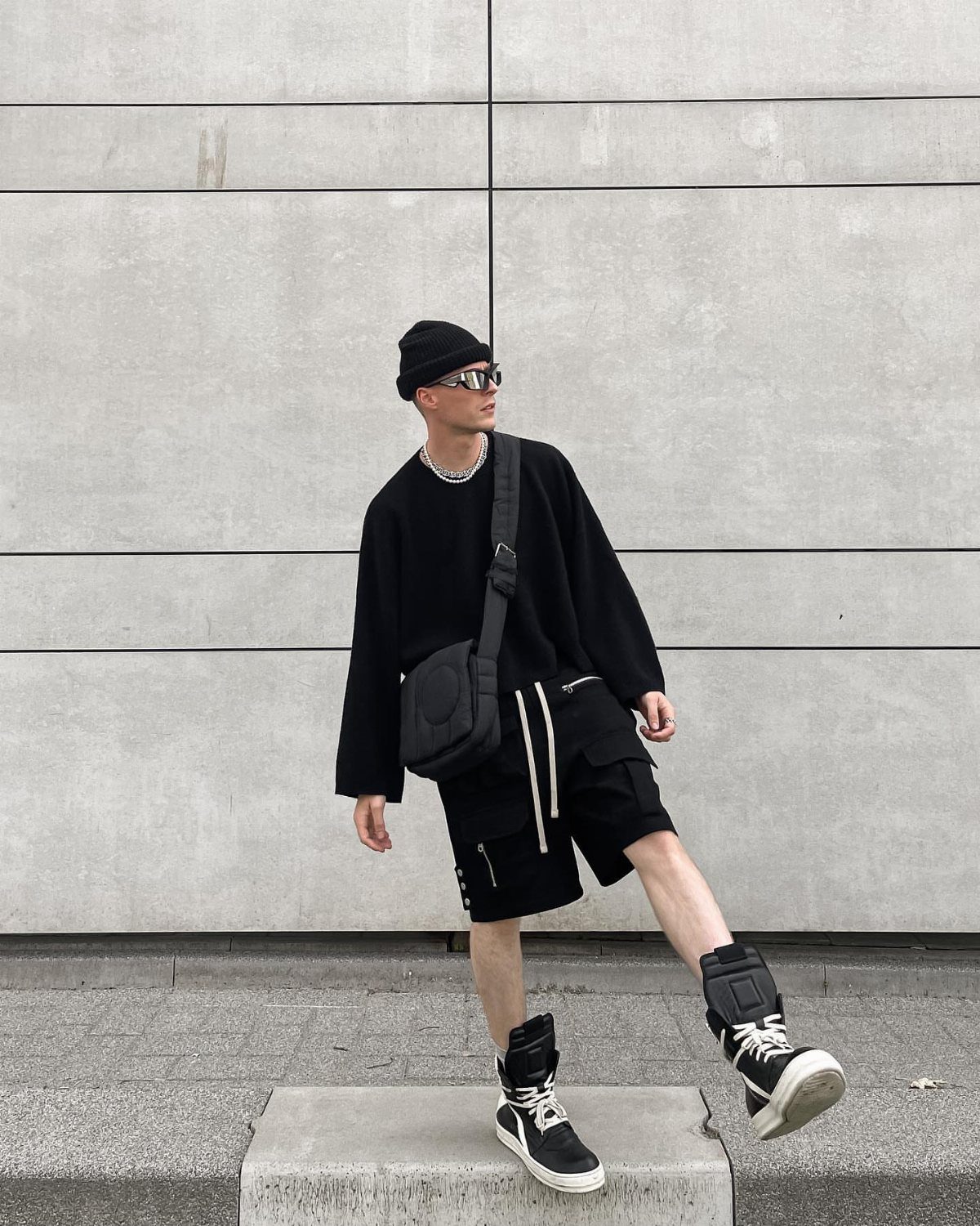 Man in oversized black streetwear and high-top sneakers posing against a concrete wall