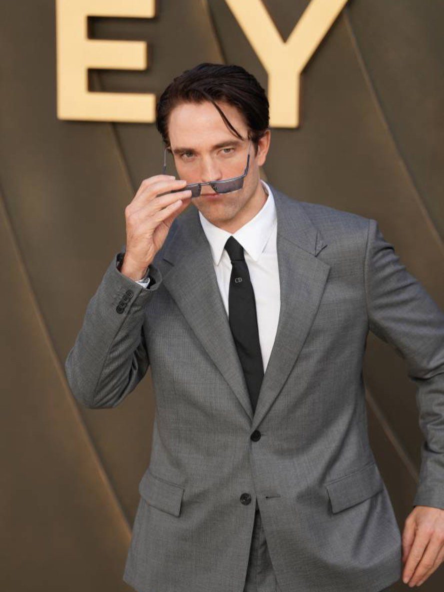 Man in a grey suit and black tie holding glasses on a red carpet.