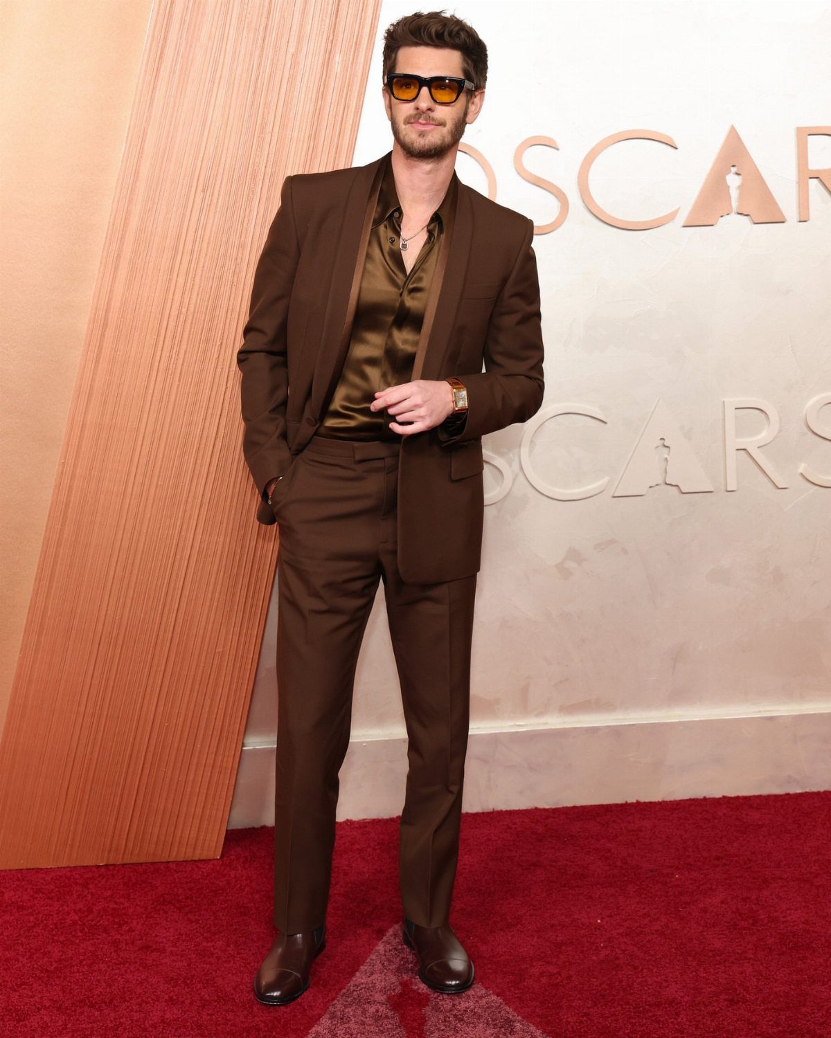 Man in a tailored brown suit and satin shirt posing on the Oscars red carpet.