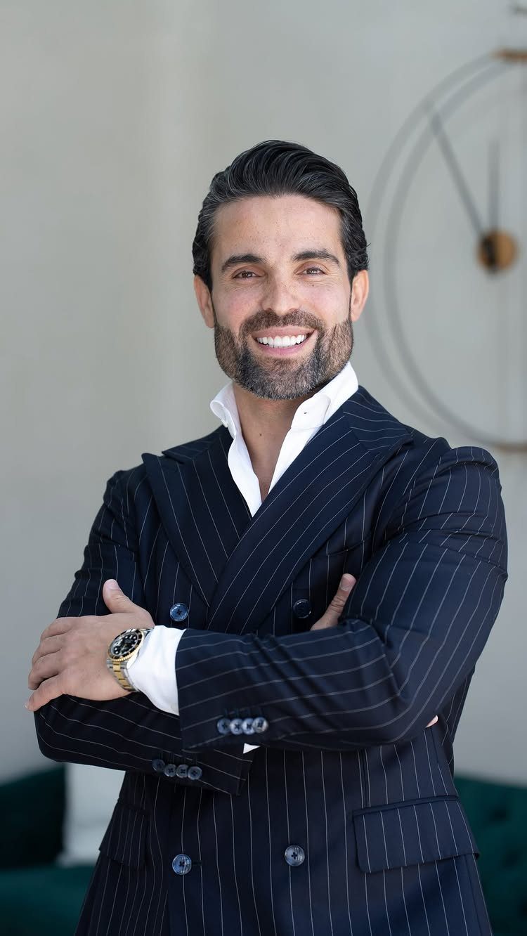 Man with slicked-back dark hair and groomed beard wearing a pinstripe navy blazer.