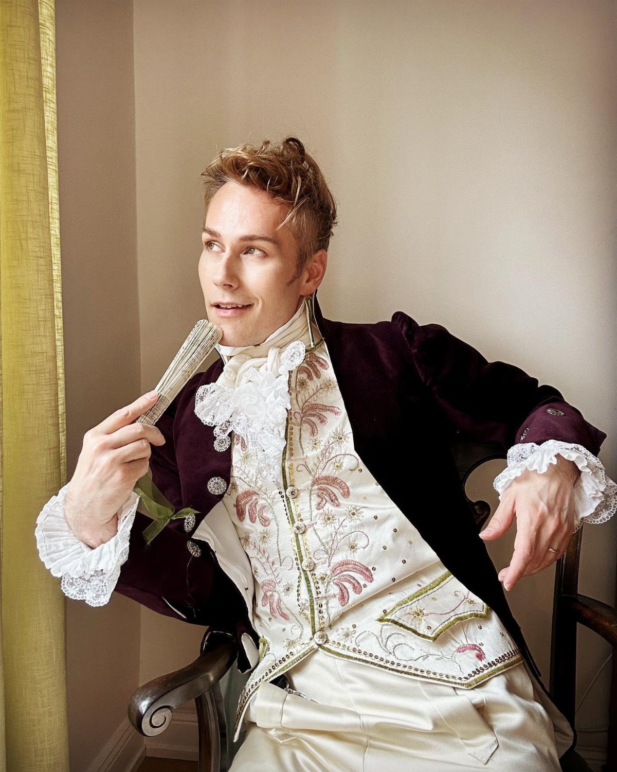 Man in regency style purple velvet coat and embroidered cream waistcoat holding a fan.