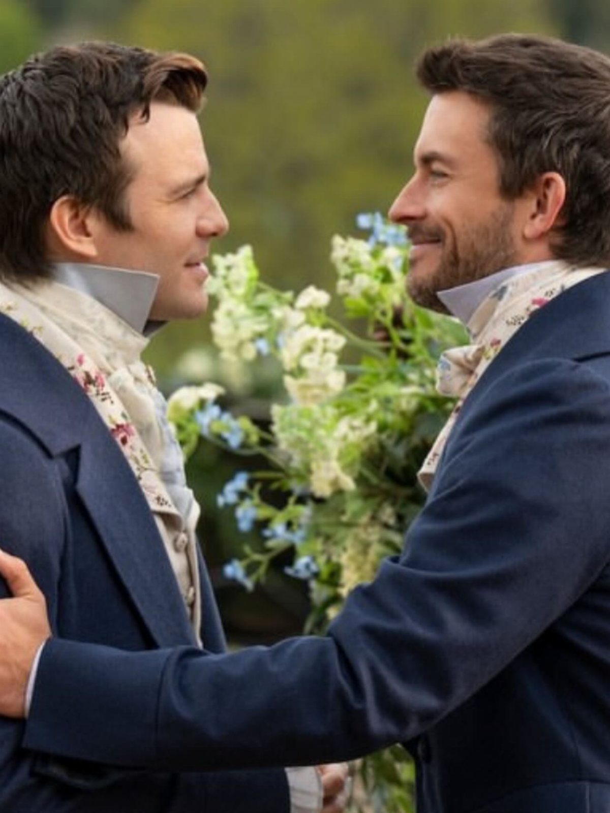 Two men in navy regency tailcoats and floral cravats facing each other outdoors