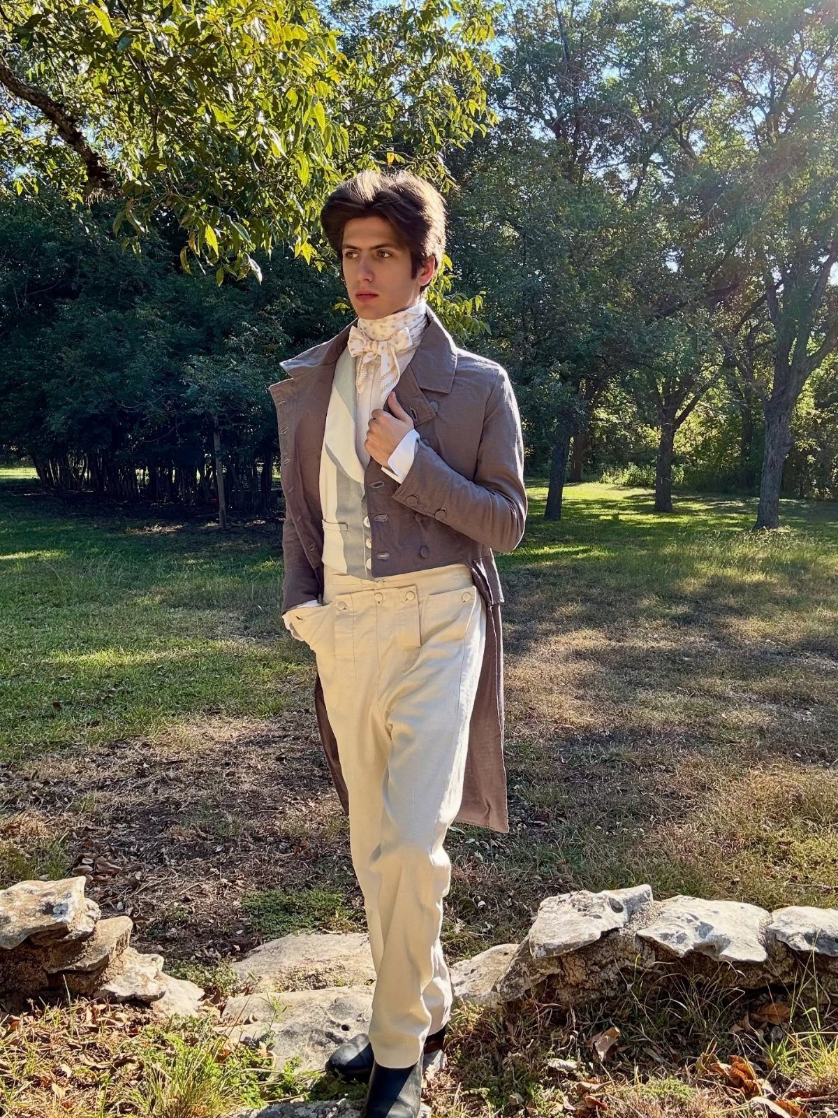 Young man wearing a brown tailcoat, white cravat, and cream trousers in a forest.