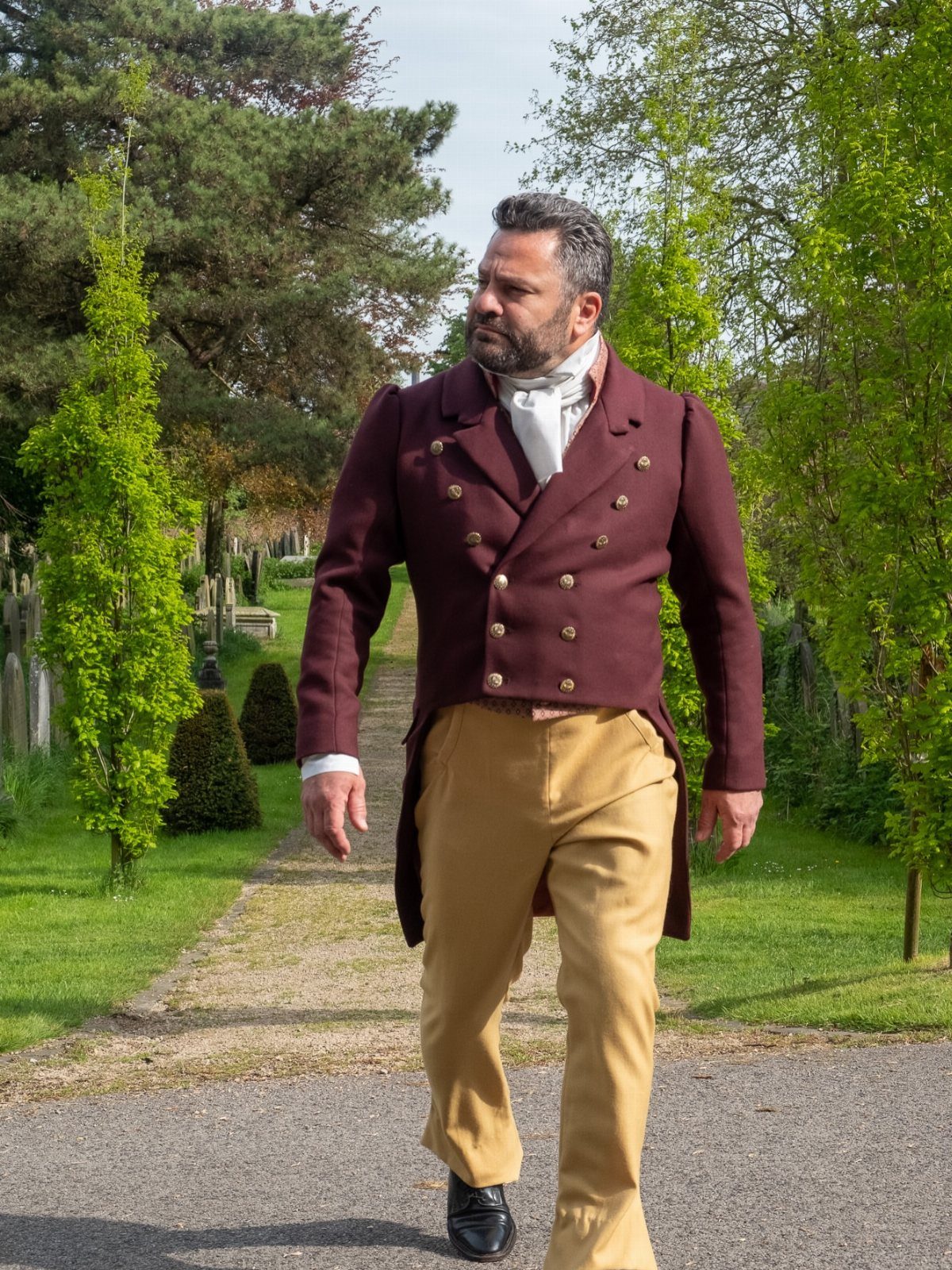 Man in maroon regency tailcoat and tan trousers walking on a garden path.