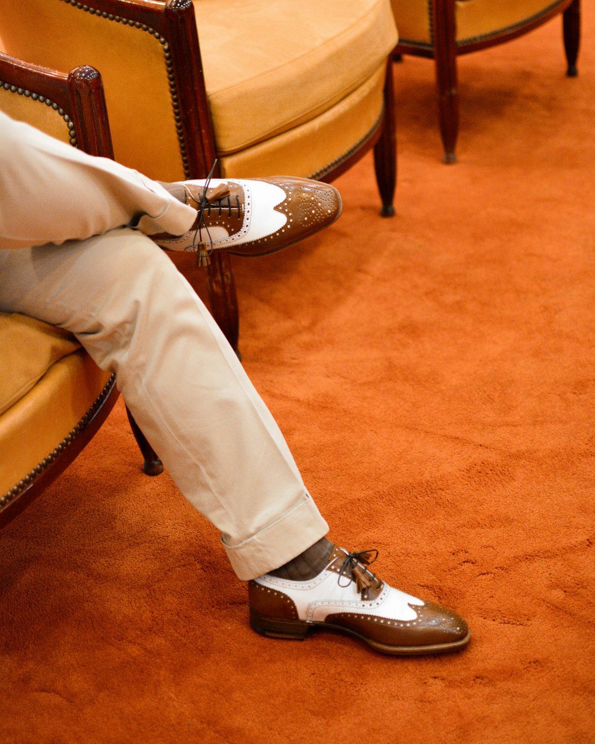 Two-tone brown and white wingtip Oxford shoes worn with beige trousers on orange carpet.