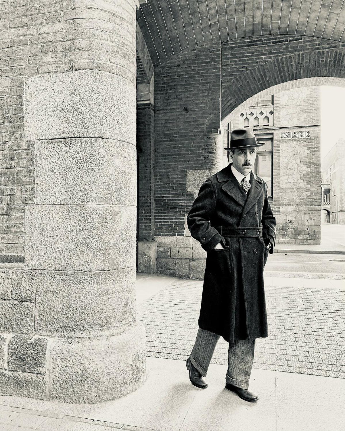 Man in vintage 1920s attire with overcoat and fedora standing by stone archway