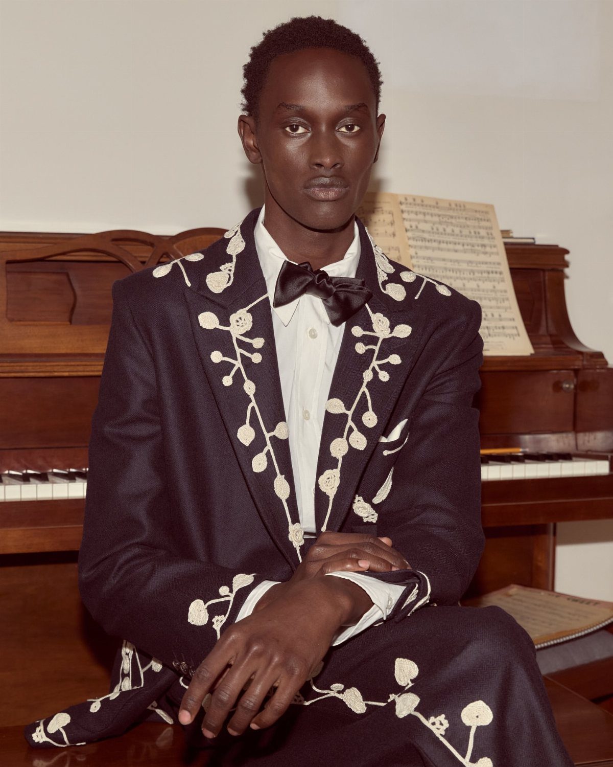 Man in a black suit with white floral embroidery and a black bow tie.