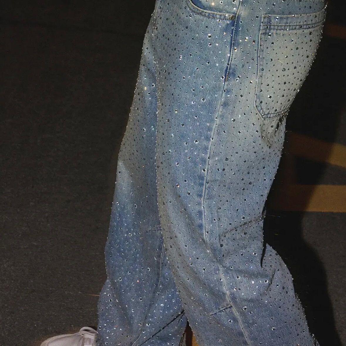Light blue denim jeans embellished with numerous sparkling silver rhinestones on dark pavement.