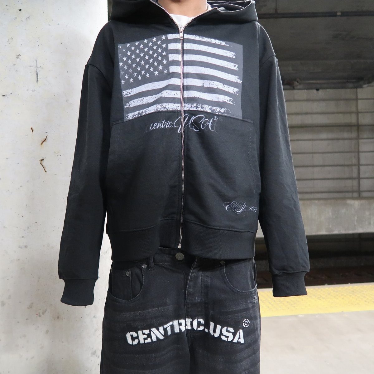 Person wearing a black oversized zip-up hoodie with American flag graphic and black jeans.