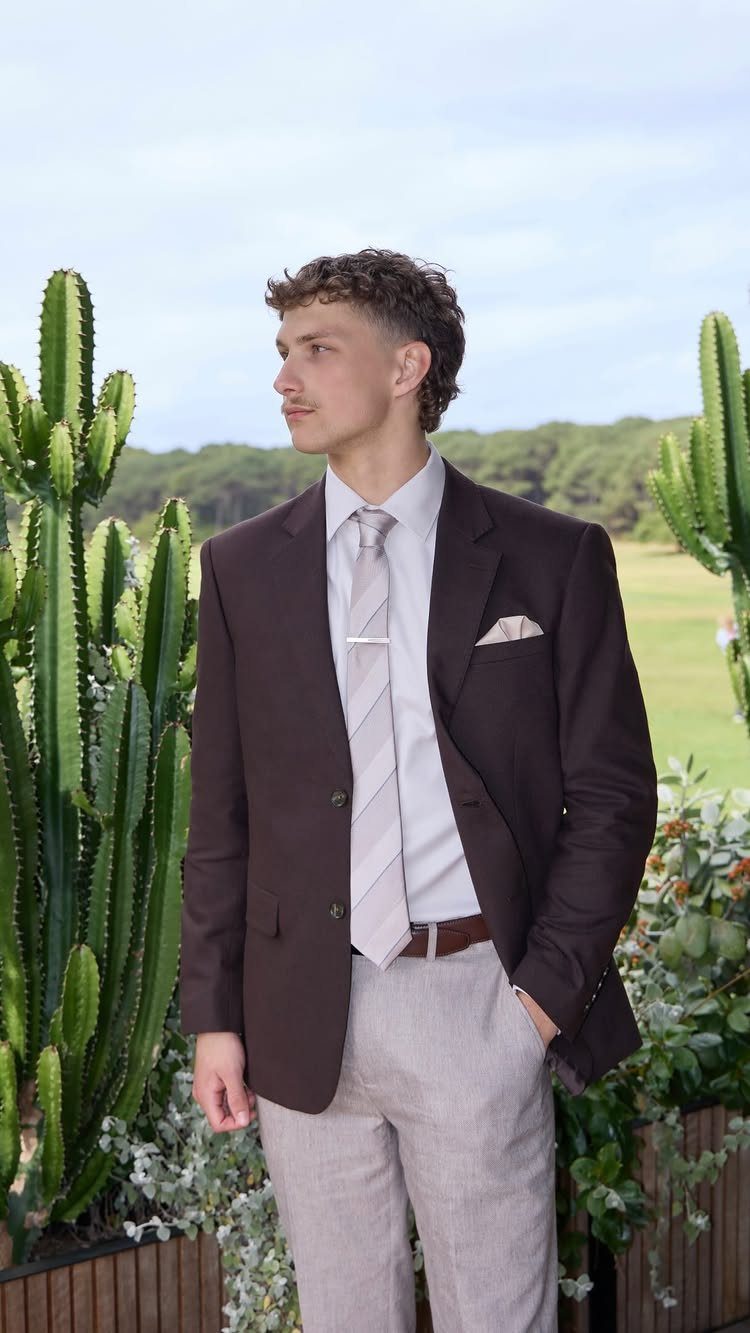 Man in a brown blazer and beige trousers posing next to tall cacti.
