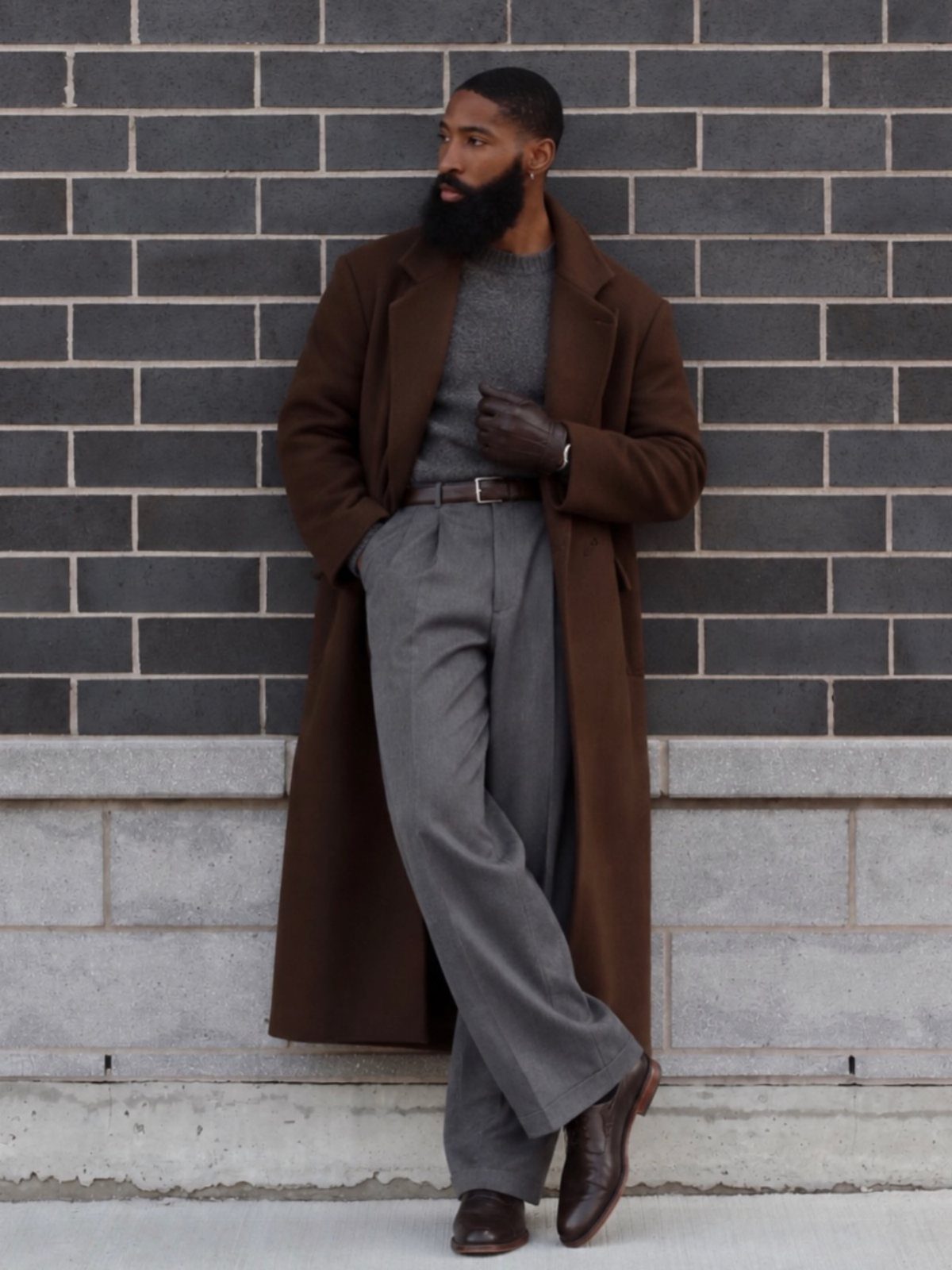 Man in a brown overcoat and grey sweater leaning against a brick wall.