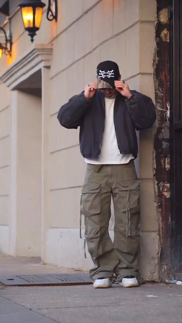 Person wearing olive green cargo pants, black jacket, and black cap against a wall.