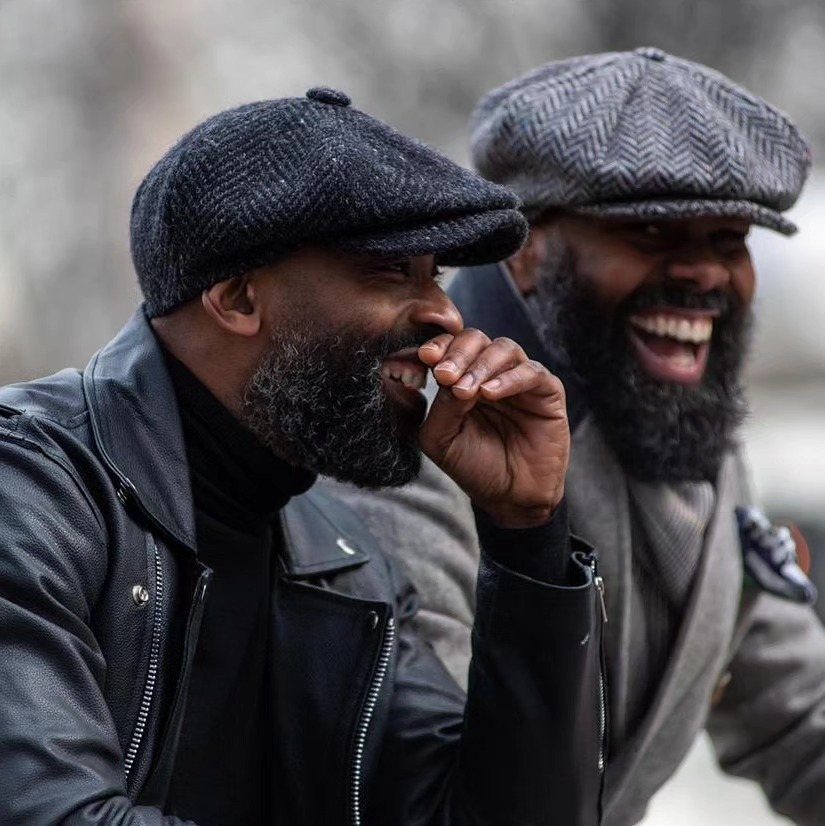 Two men wearing flat caps and stylish winter jackets laughing together outdoors.