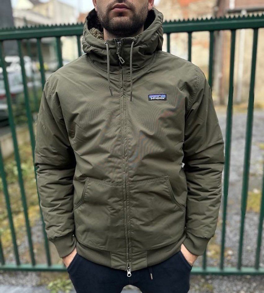 Man wearing an olive green Patagonia hooded jacket standing before a green fence.