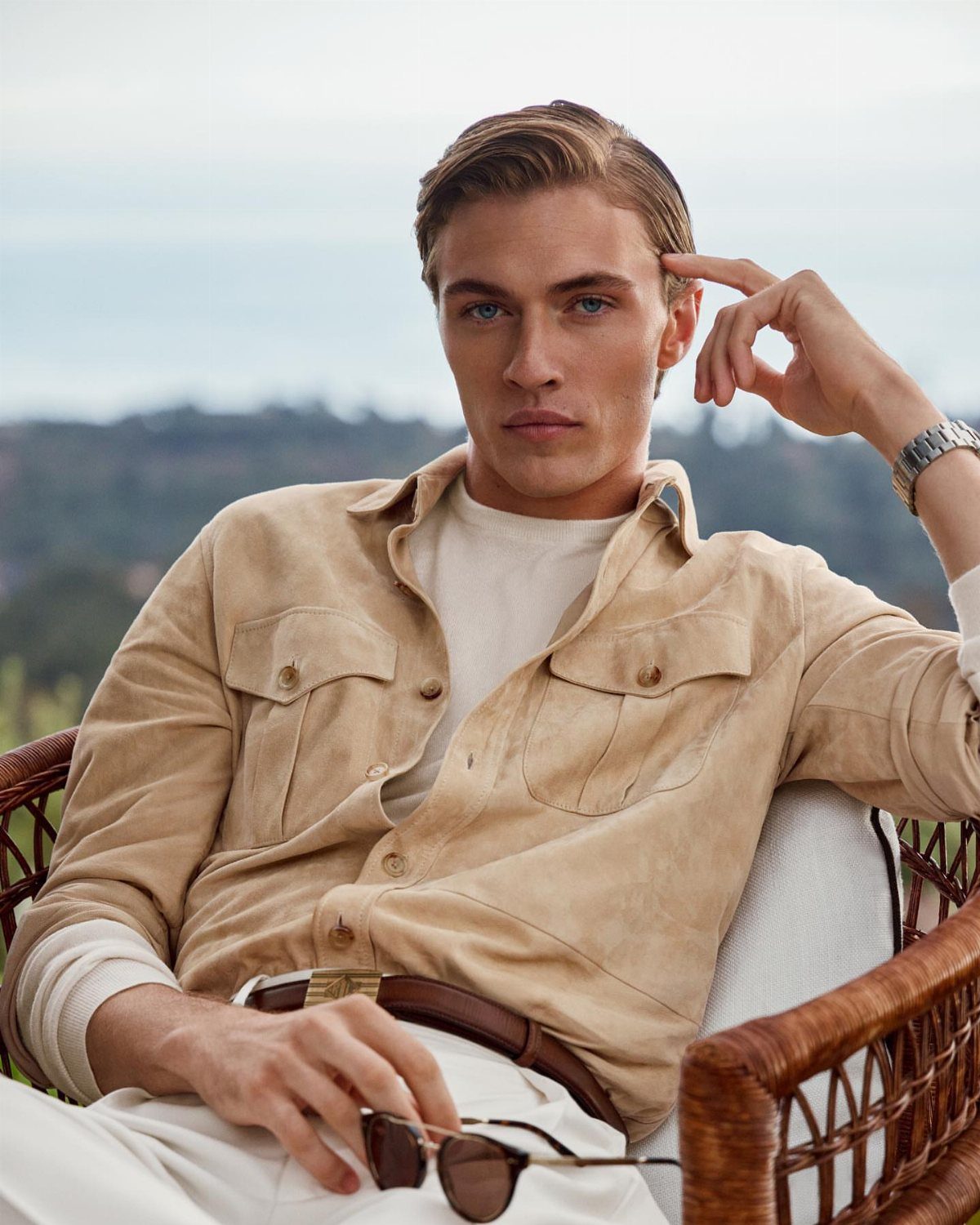 Man in tan suede shirt and white trousers sitting in a wicker chair.