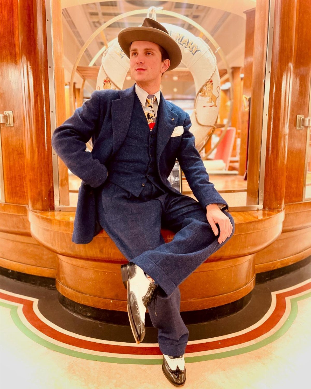Man in a navy blue three-piece tailored suit and brown hat sitting indoors.