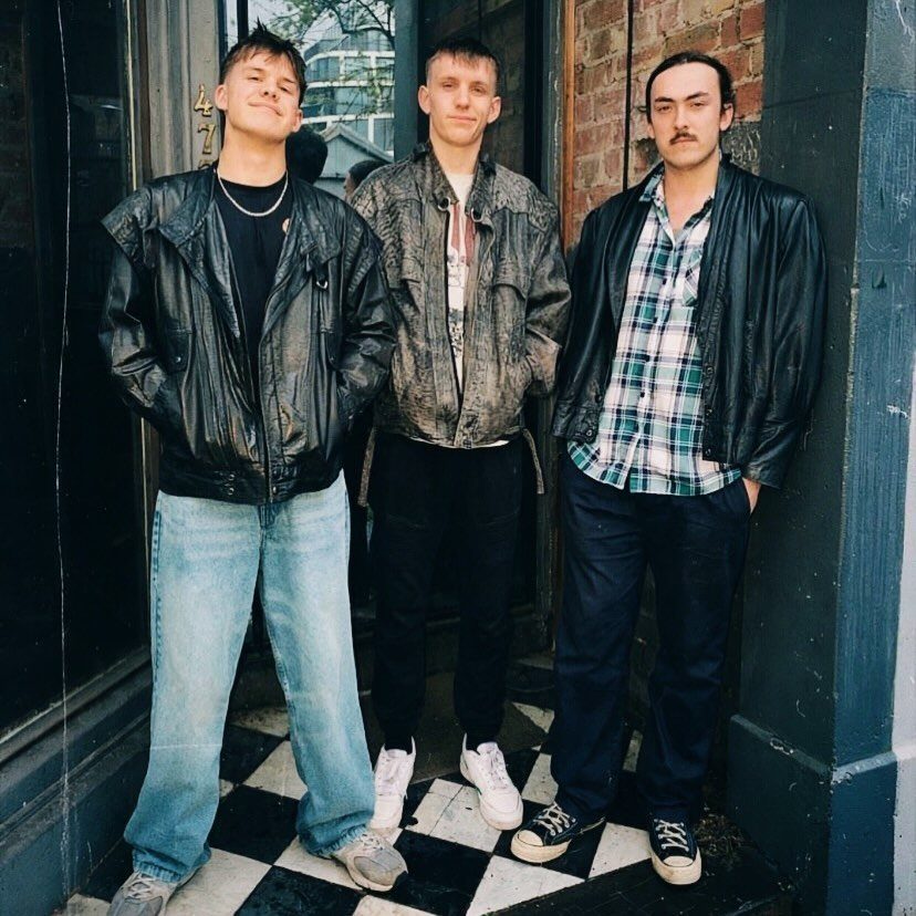 Three men wearing vintage leather jackets and jeans standing in front of a building.