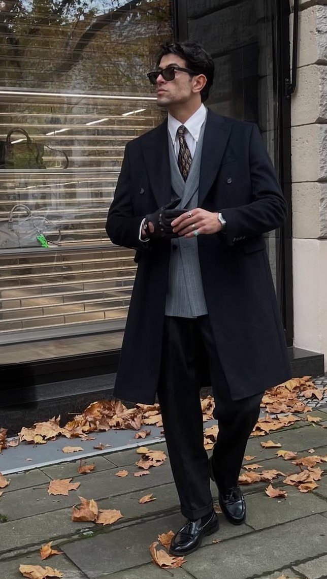 Man in a black overcoat and grey waistcoat standing on autumn leaves.